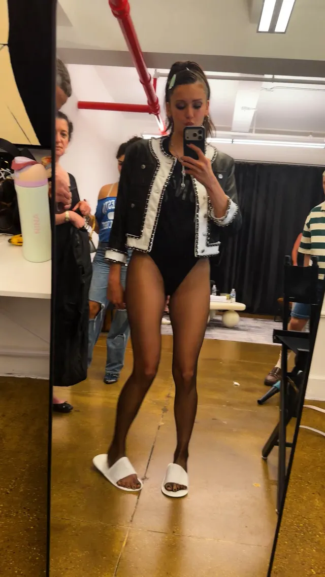Nina Dobrev taking a mirror photo wearing a jacket and white slides.