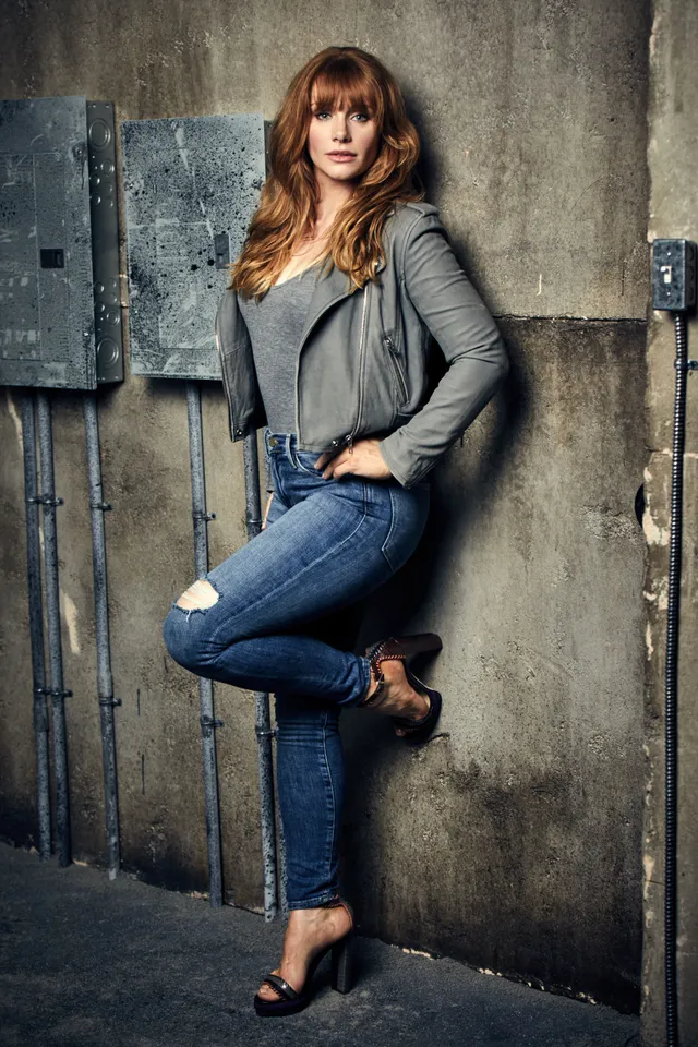 Bryce Dallas Howard posing beside a wall in jeans and a grey jacket.