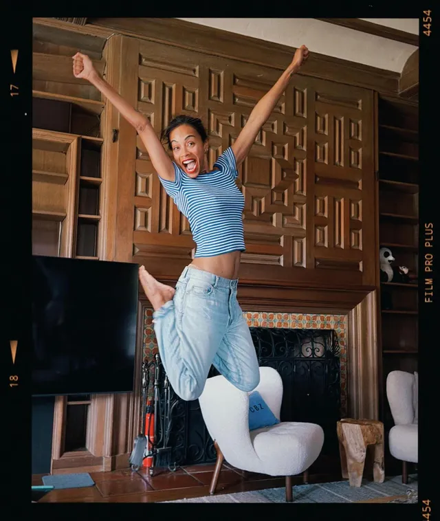 Zoe Saldaña jumping indoors in jeans and a blue top.
