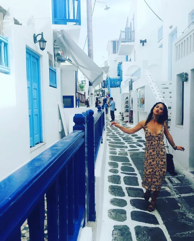 Adria Arjona walking along a narrow street in a patterned dress.