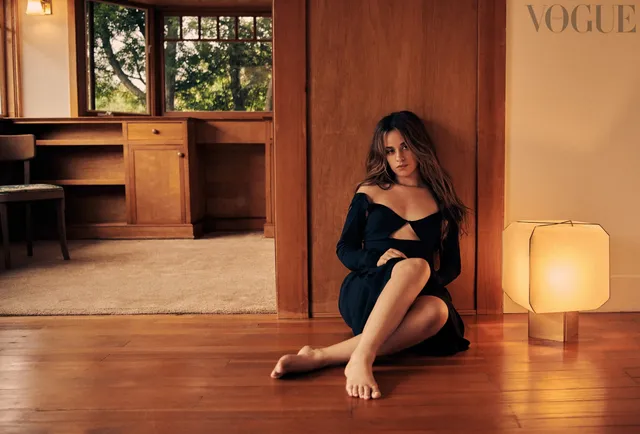 Camila Cabello sitting barefoot on a wooden floor in a dark dress.
