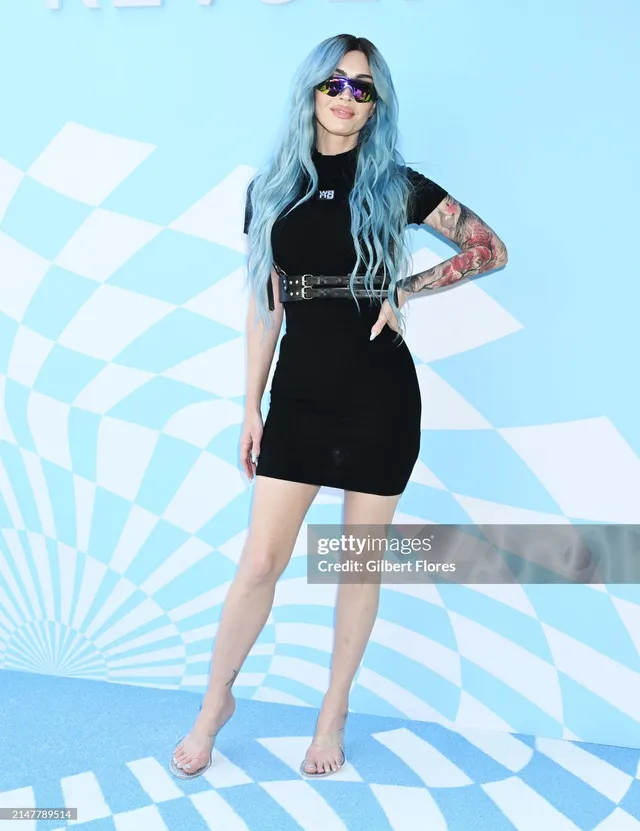Megan Fox posing in a black dress and pale heels beside a blue backdrop.