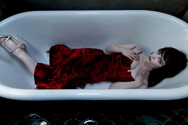 Sophie Thatcher reclining in a bathtub in a red dress and metallic heels.