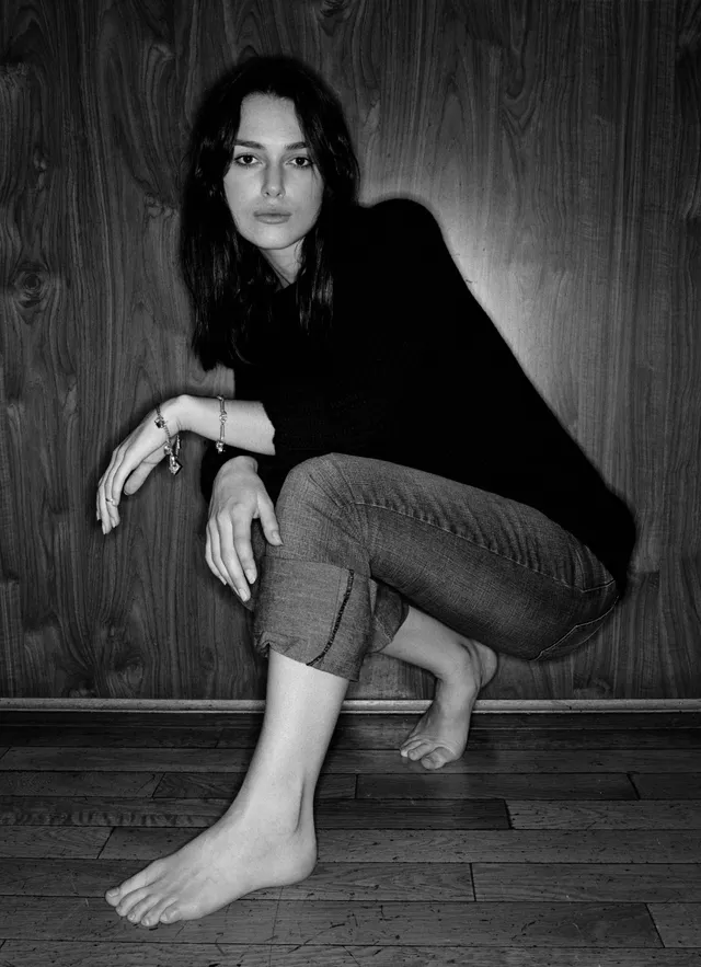 Keira Knightley a monochrome crouching portrait beside a wooden wall.