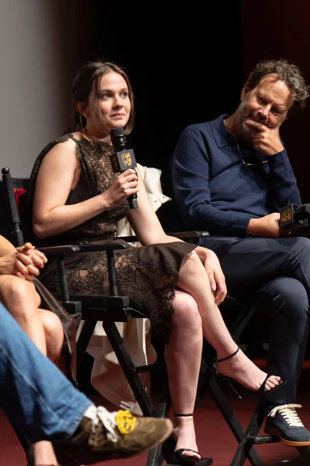 Cailee Spaeny sitting at a panel discussion in a dark dress and black heels.