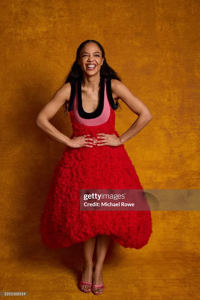 Tessa Thompson posing in a red dress against a gold studio background.