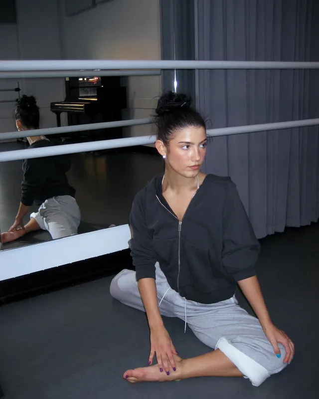 Charli D’Amelio sitting beside a dance studio mirror with one bare foot.