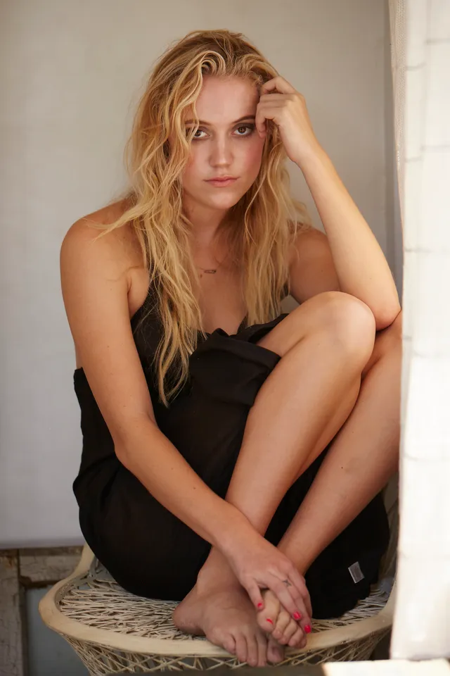 Maika Monroe sitting barefoot on a wicker stool.