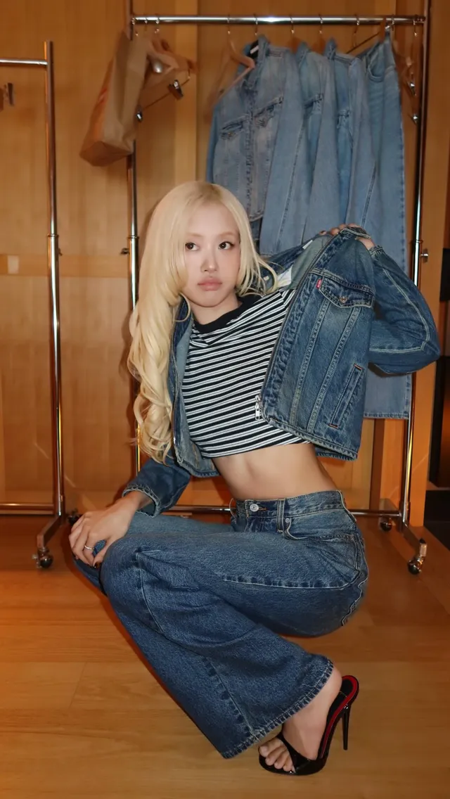 Rosé crouching beside hanging clothes in a striped top, jeans and black heels.