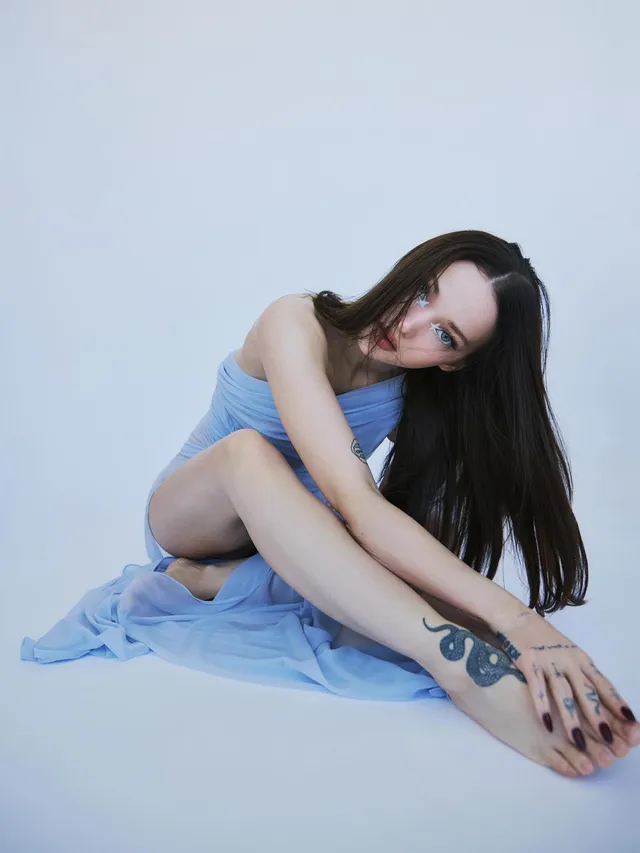 Dove Cameron sitting barefoot in a pale blue studio dress.