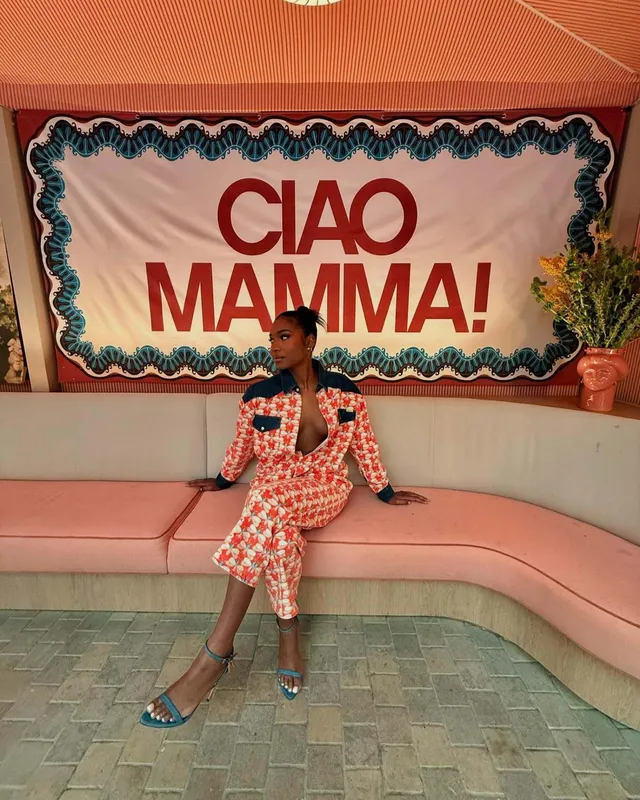Carlacia Grant sitting on a pink sofa in a patterned suit and blue heels.