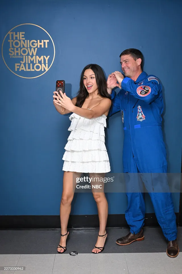 Olivia Rodrigo posing beside a man in a white dress and black heels.