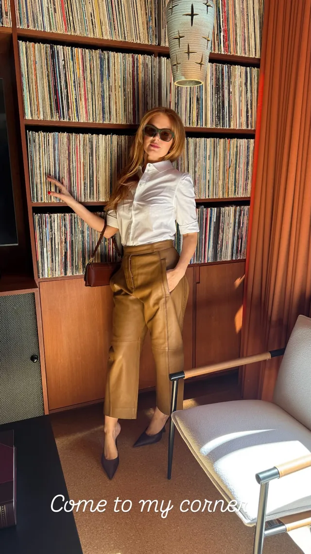 Isla Fisher posing beside shelves in a pale blouse and gold trousers.