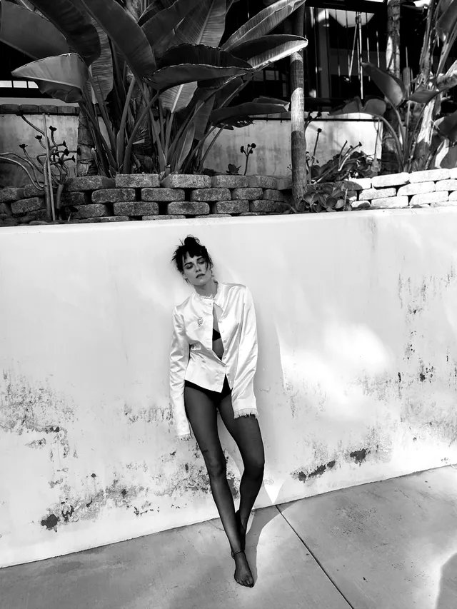 Kristen Stewart posing barefoot beside a wall in a monochrome portrait.