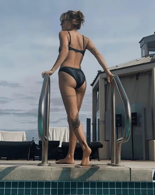 Ella Purnell standing at a pool ladder in a black swimsuit.