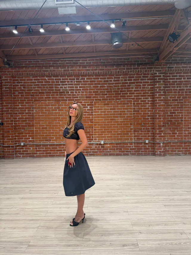 Ester Expósito standing in black heeled mules and a dark skirt during a studio scene.
