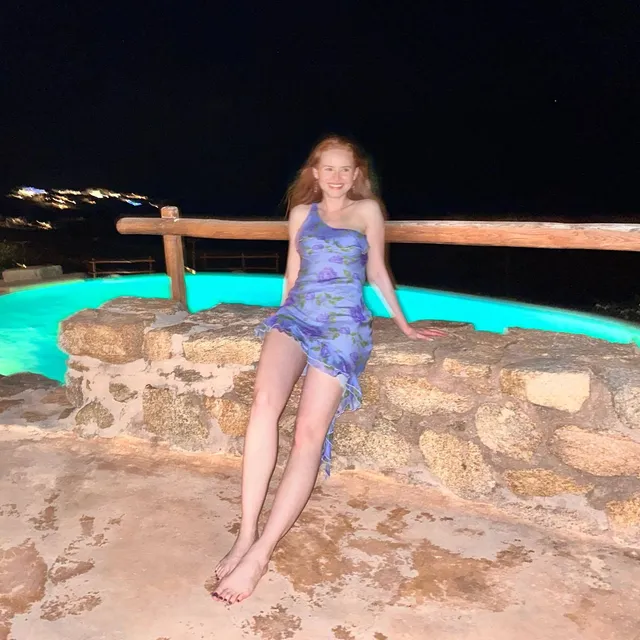 Madelaine Petsch sitting barefoot on a stone wall beside a pool at night.