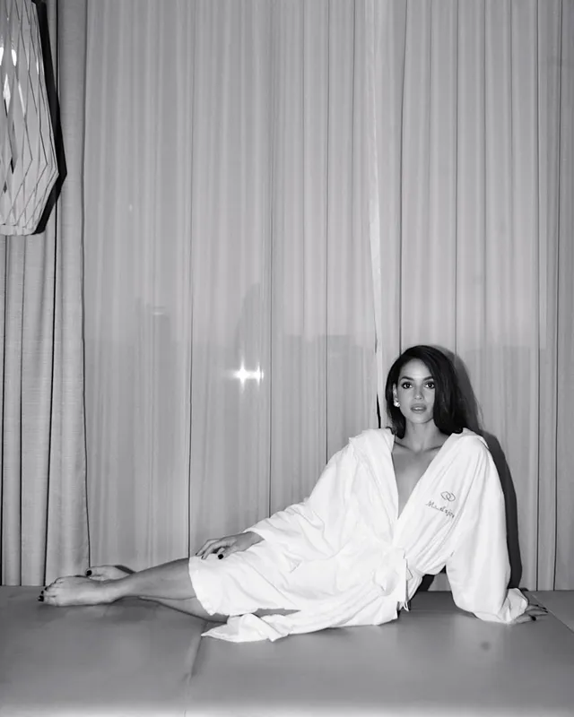 Adria Arjona sitting barefoot on a studio floor in a white robe.