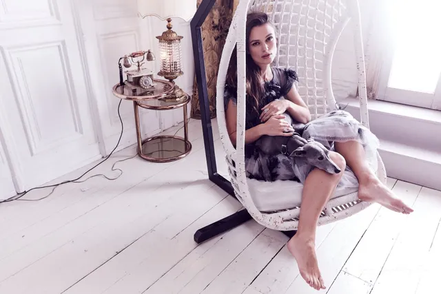 Keira Knightley seated in a white hanging chair in a softly lit room.
