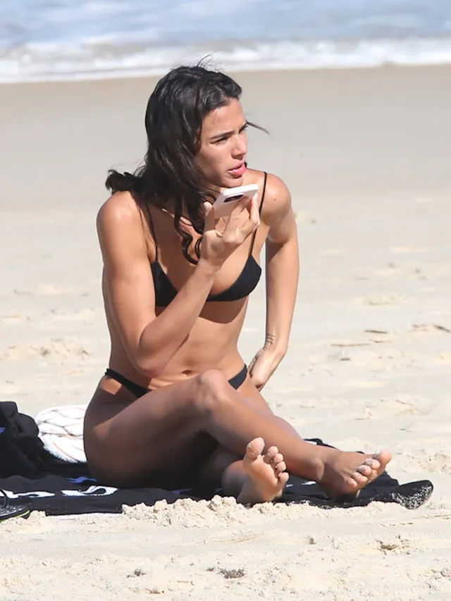 Bruna Marquezine sitting barefoot on a beach in a black bikini.