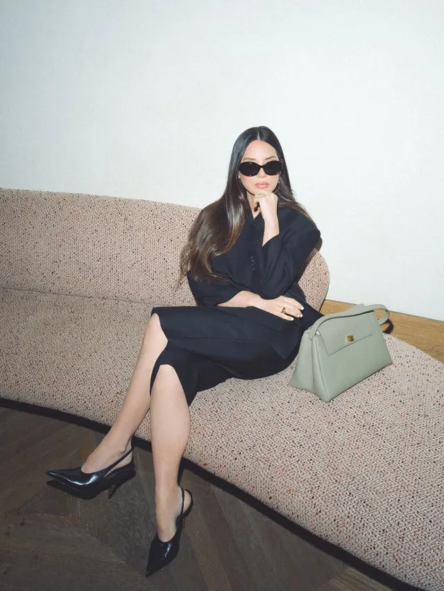 Olivia Munn sitting in a black dress and pointed heels.