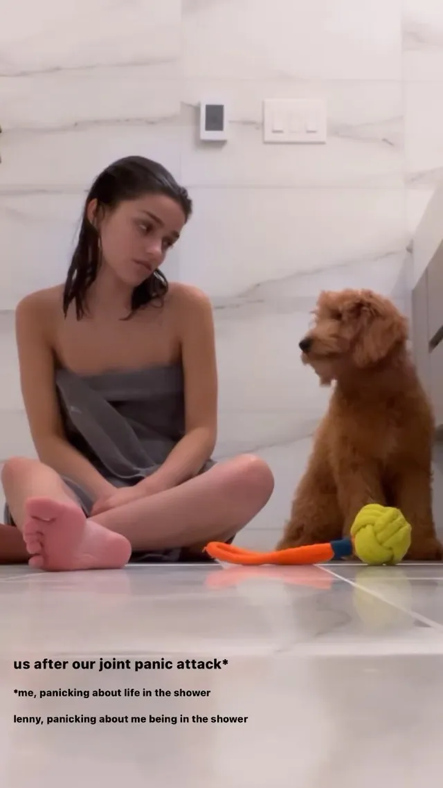 Rachel Zegler sitting barefoot on the floor beside a dog.