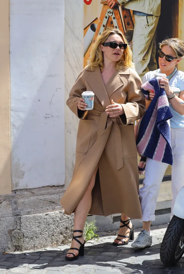 Florence Pugh walking with a companion in a tan coat and strappy heels.