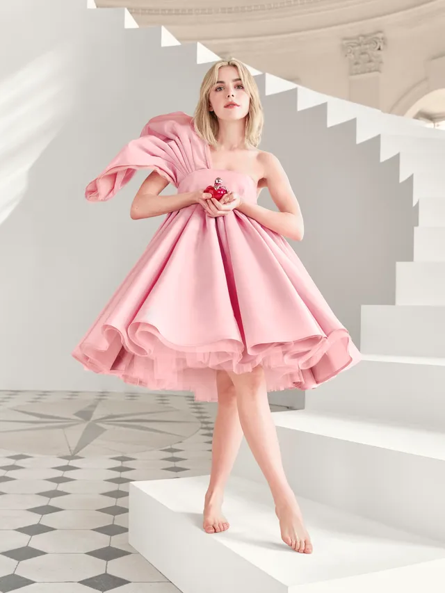 Kiernan Shipka standing barefoot beside a white staircase in a pink dress.