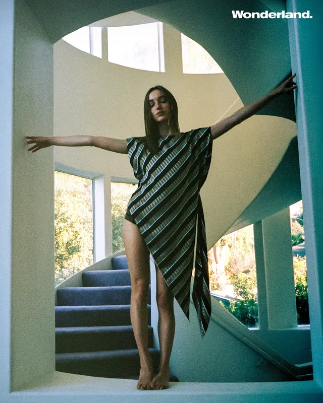 Phoebe Dynevor standing barefoot on a spiral staircase in a striped dress.