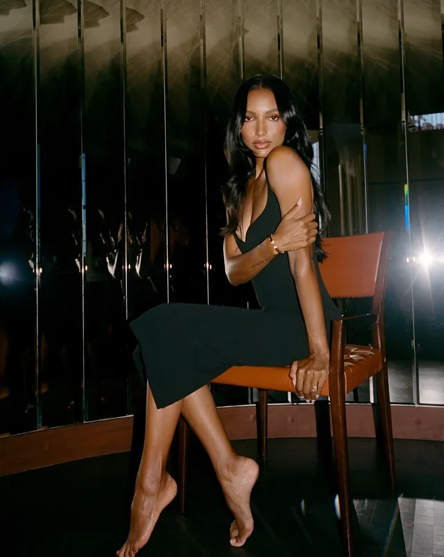 Jasmine Tookes sitting in a chair wearing heels and a dark green dress.
