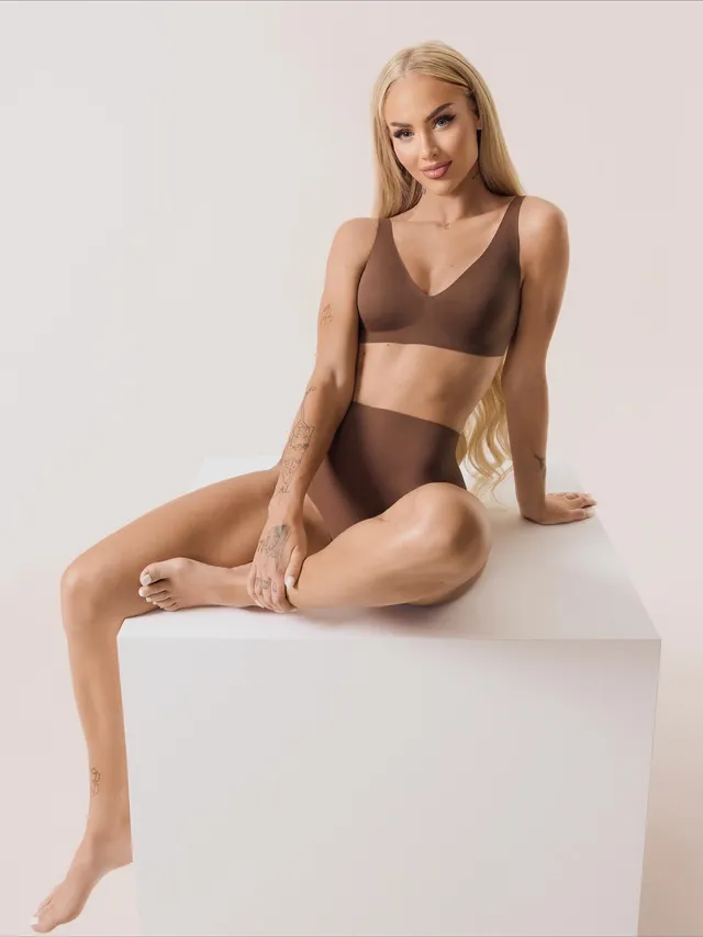 Alisha Lehmann sitting barefoot on a studio pedestal in a brown outfit.