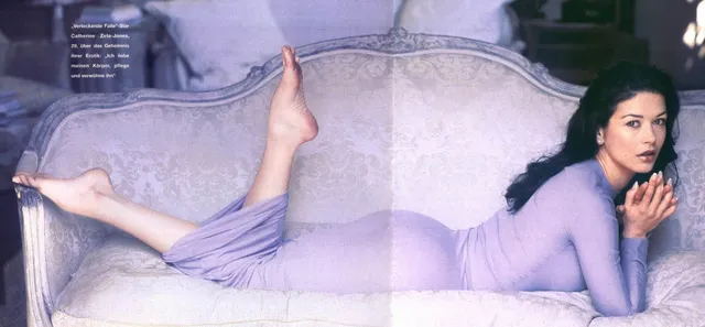 Catherine Zeta-Jones reclining on a pale sofa in a lavender outfit.