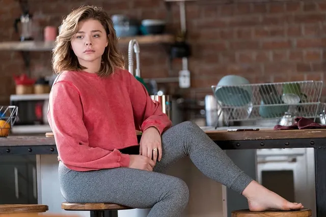 Chloë Grace Moretz sitting on a kitchen counter in gray trousers with a bare foot extended.