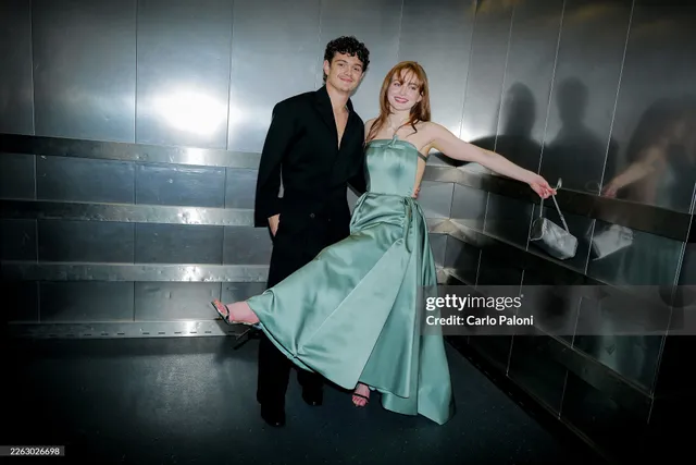 Sadie Sink posing beside a man on stairs in a green gown and heels.