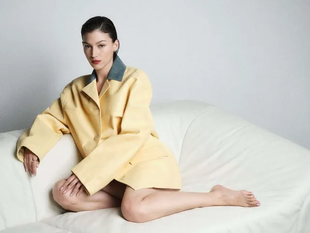Úrsula Corberó sitting barefoot on a pale sofa in a yellow jacket.