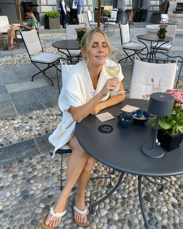 Claire Holt sitting at an outdoor cafe table in a white outfit and flip-flops.