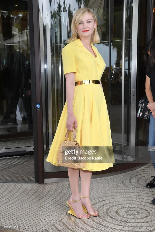 Kirsten Dunst posing outside in a yellow dress and matching heels.