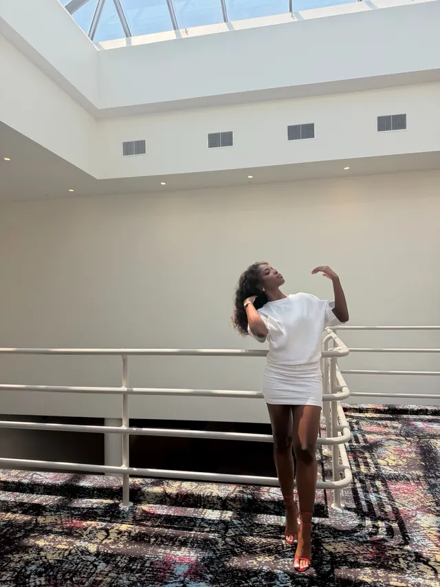 Ryan Destiny posing in a white shirt-style dress and heels indoors.