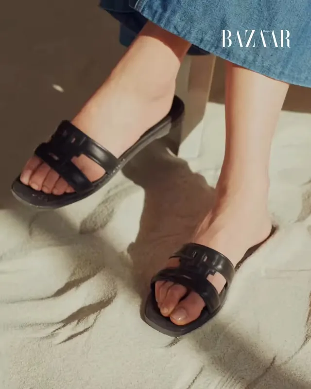 Jisoo showing black flat slides on a sandy surface during a fashion shoot.