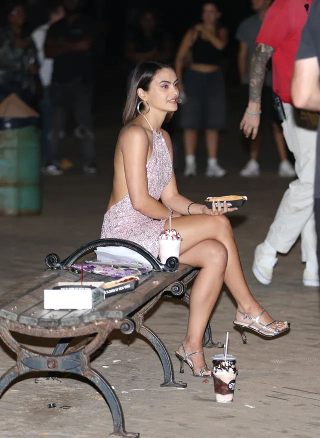 Camila Mendes sitting on a bench in a pink dress and heels.