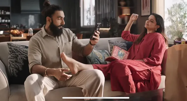 Deepika Padukone reclining on a sofa in a red dress with her bare foot raised.