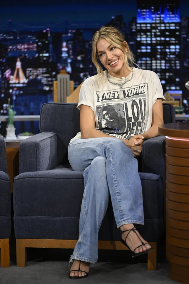 Sienna Miller sitting on a talk show sofa in jeans.