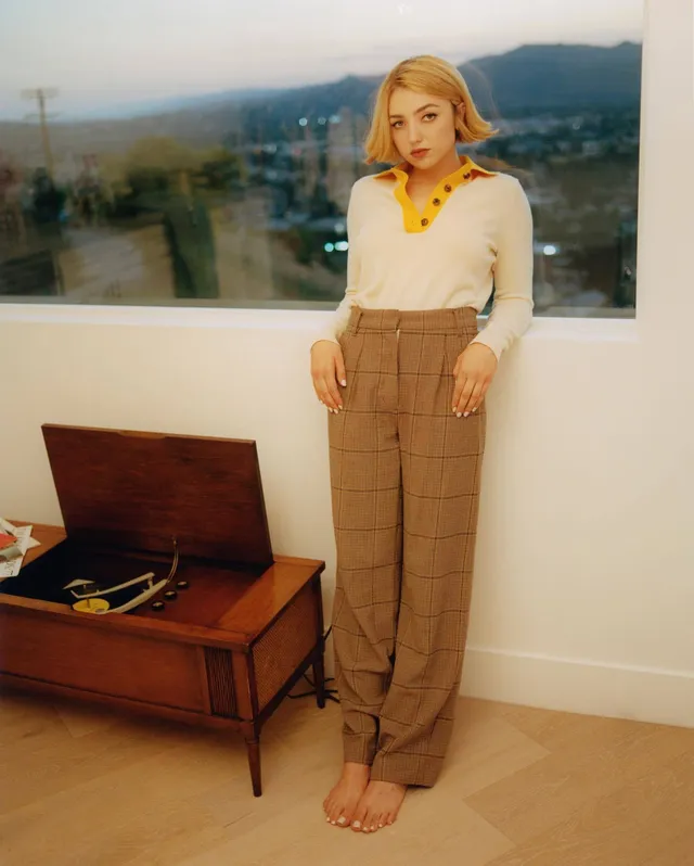 Peyton List standing barefoot in brown trousers indoors.