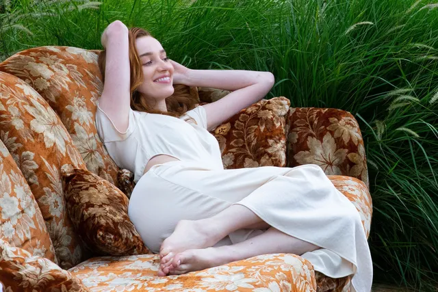 Phoebe Dynevor relaxing on a floral armchair in a white dress.