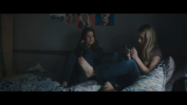 Grace Van Patten sitting barefoot beside another woman on a bed in a film scene.