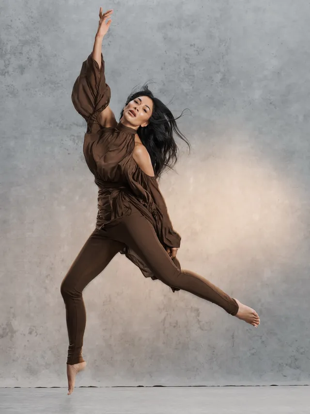 Nicole Scherzinger balancing barefoot in a brown outfit in a studio photograph.