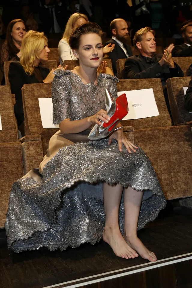 Kristen Stewart sitting barefoot at an event holding her high heels.