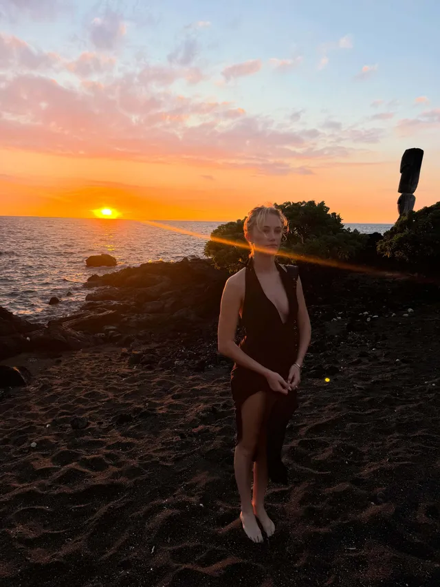 Maika Monroe standing on rocks beside the sea at sunset.