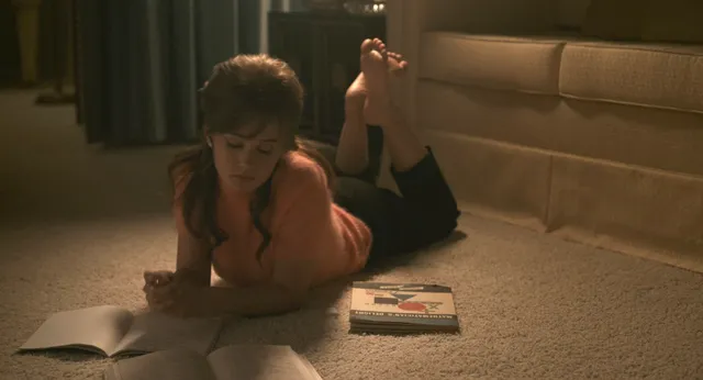 Cailee Spaeny reading on a bedroom floor with her bare feet raised.