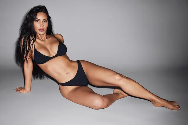 Kim Kardashian: reclining barefoot in a black outfit against a grey background.